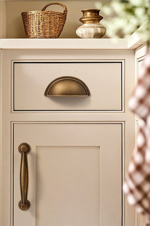 Solid brass beaded-rim half-moon cup pull in Antique Brass on a cream cabinet drawer.