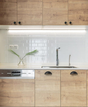 Solid brass open-back cup pulls in Blackened Bronze shown on wood-look kitchen cabinets below a sink.