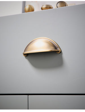 Solid brass open-back cup pull in Burnished Brass on a gray drawer, viewed from below.