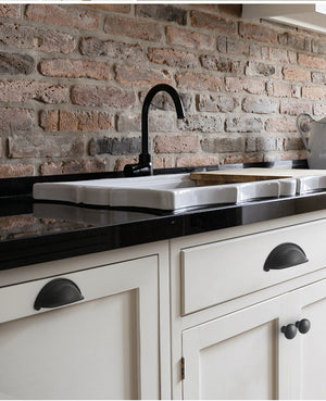 Solid brass open-back cup pulls in Matte Black shown on white kitchen fronts below a sink and brick backsplash.