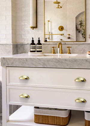 Four polished gold half-moon cup pulls on a white bathroom vanity beneath a light veined countertop.
