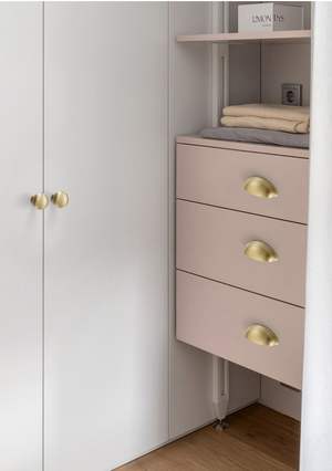 Three satin brass half-moon cup pulls on pale pink drawers beside white cabinet doors.