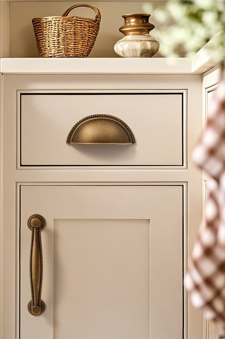 Solid brass beaded-rim half-moon cup pull in Antique Brass on a cream cabinet drawer.