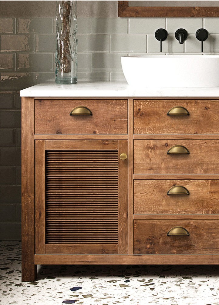 Solid brass open-back cup pulls in Aged Brass on the drawers of a wood-grain bathroom vanity.