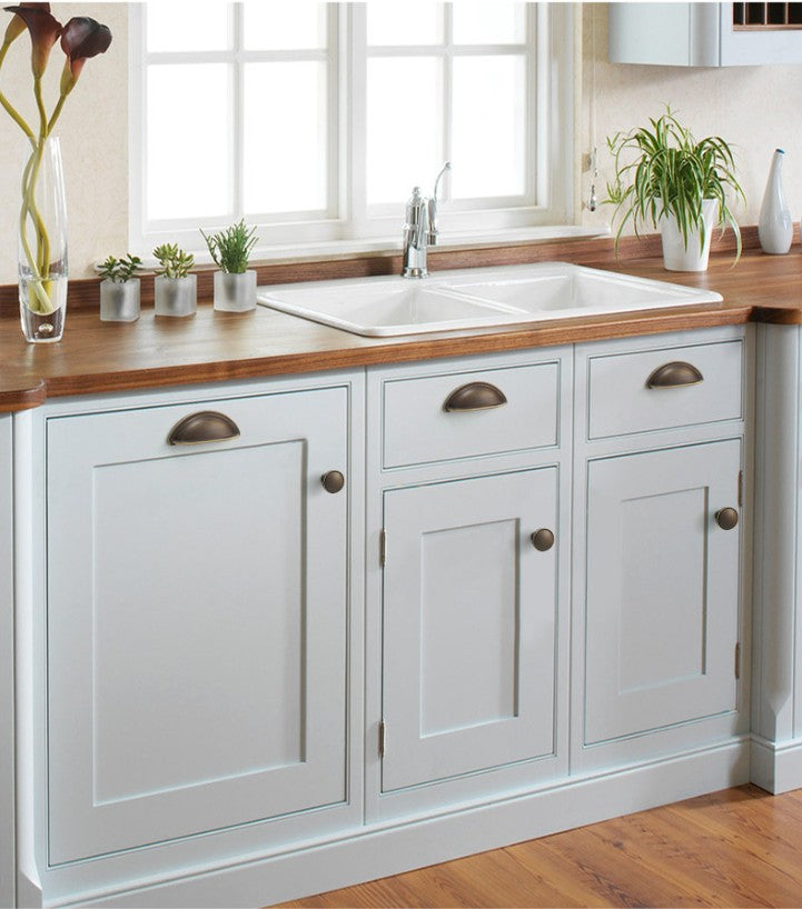 Solid brass open-back cup pulls in Antique Bronze on light-colored kitchen cabinetry beneath a sink.