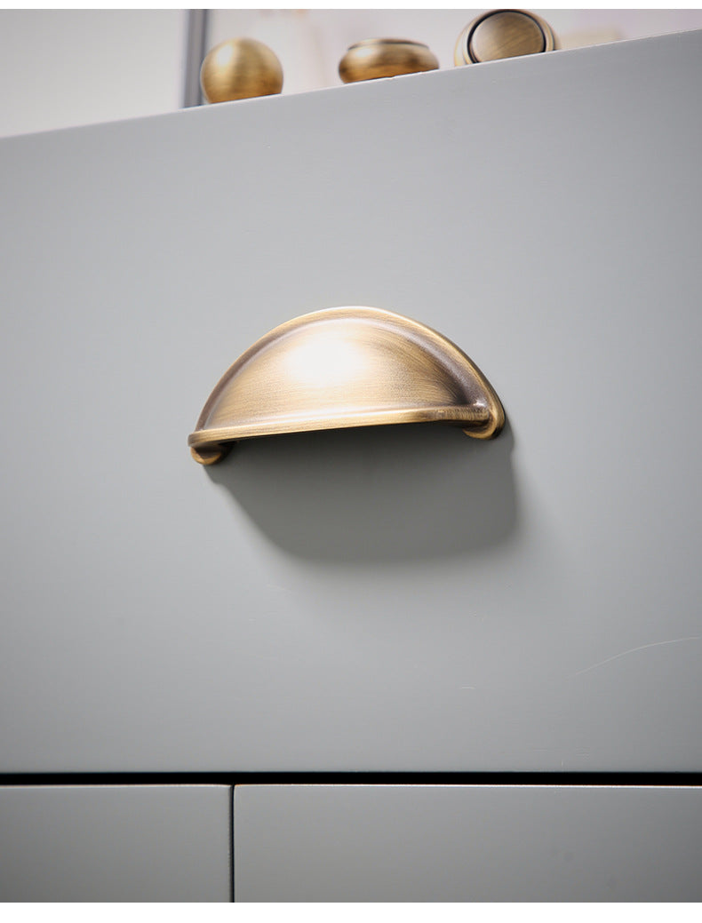 Solid brass open-back cup pull in Burnished Brass on a gray drawer, viewed from below.