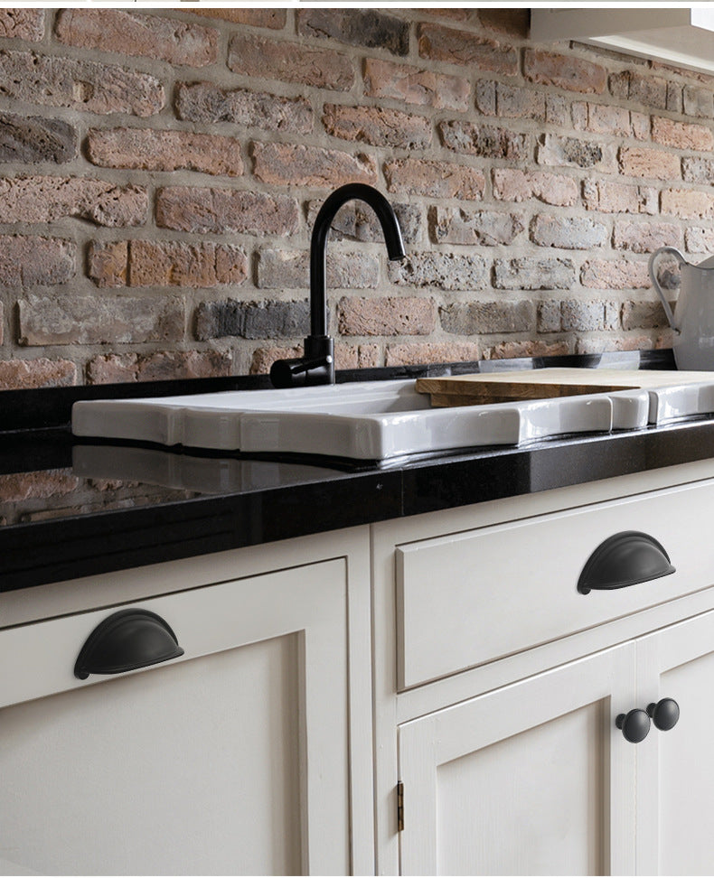 Solid brass open-back cup pulls in Matte Black shown on white kitchen fronts below a sink and brick backsplash.