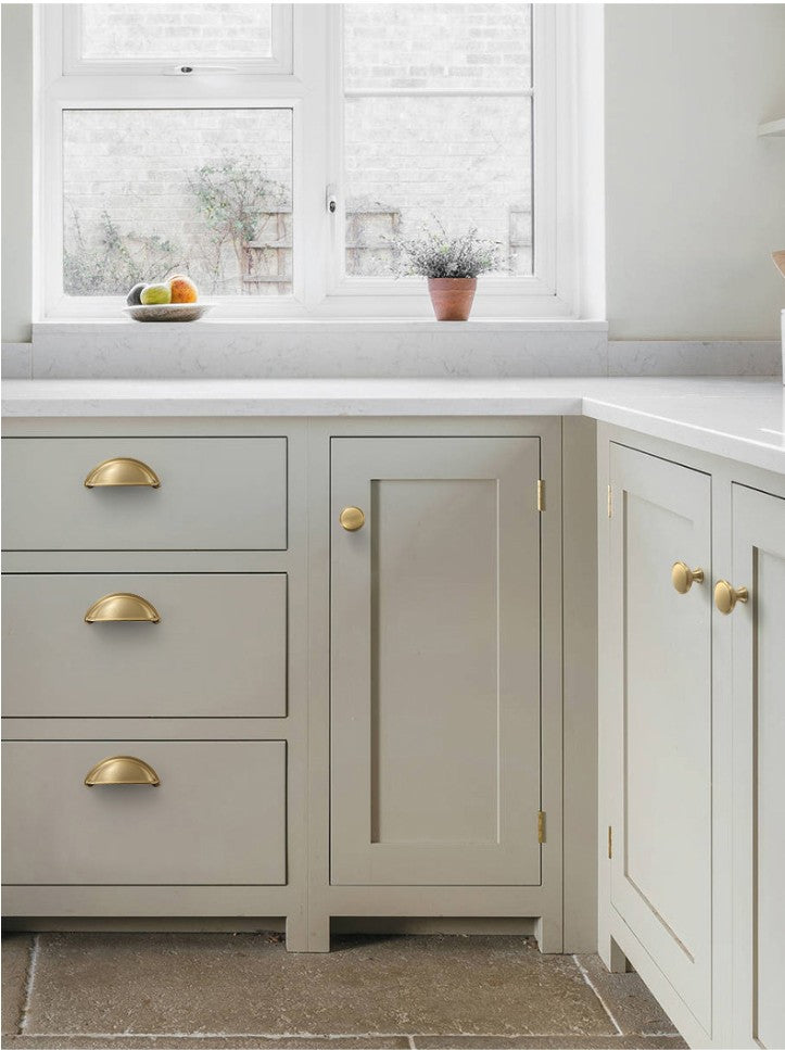 Solid brass open-back cup pulls in Satin Brass on light-colored kitchen drawers beneath a window.