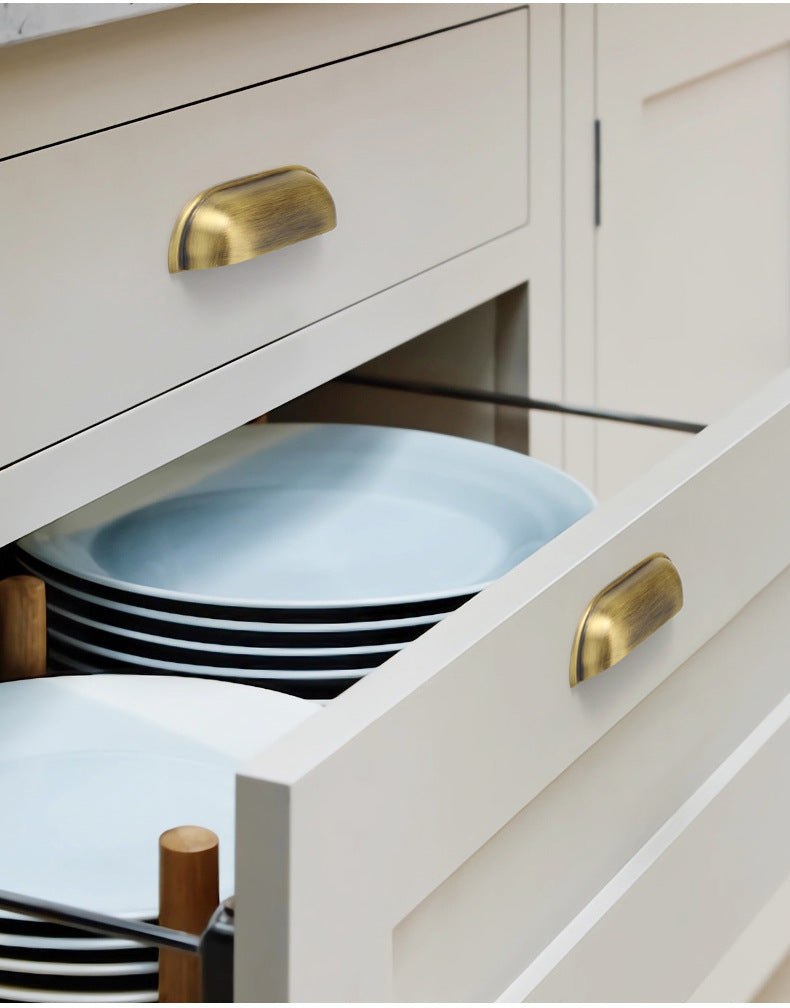 Solid brass soft-corner cup pulls in Aged Brass on cream kitchen drawers, with one drawer open.