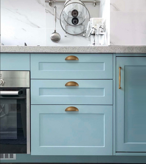 Three aged brass half-moon cup pulls on pale blue kitchen drawers.