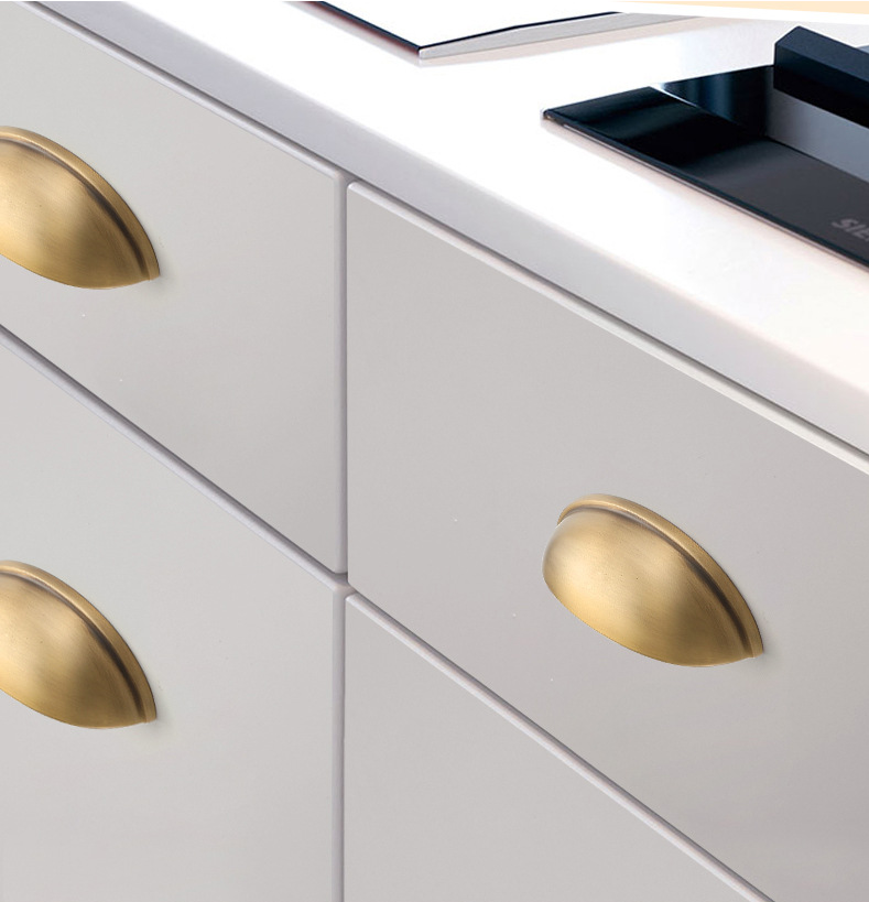 Burnished brass half-moon cup pulls mounted on pale gray kitchen drawers beside a cooktop.