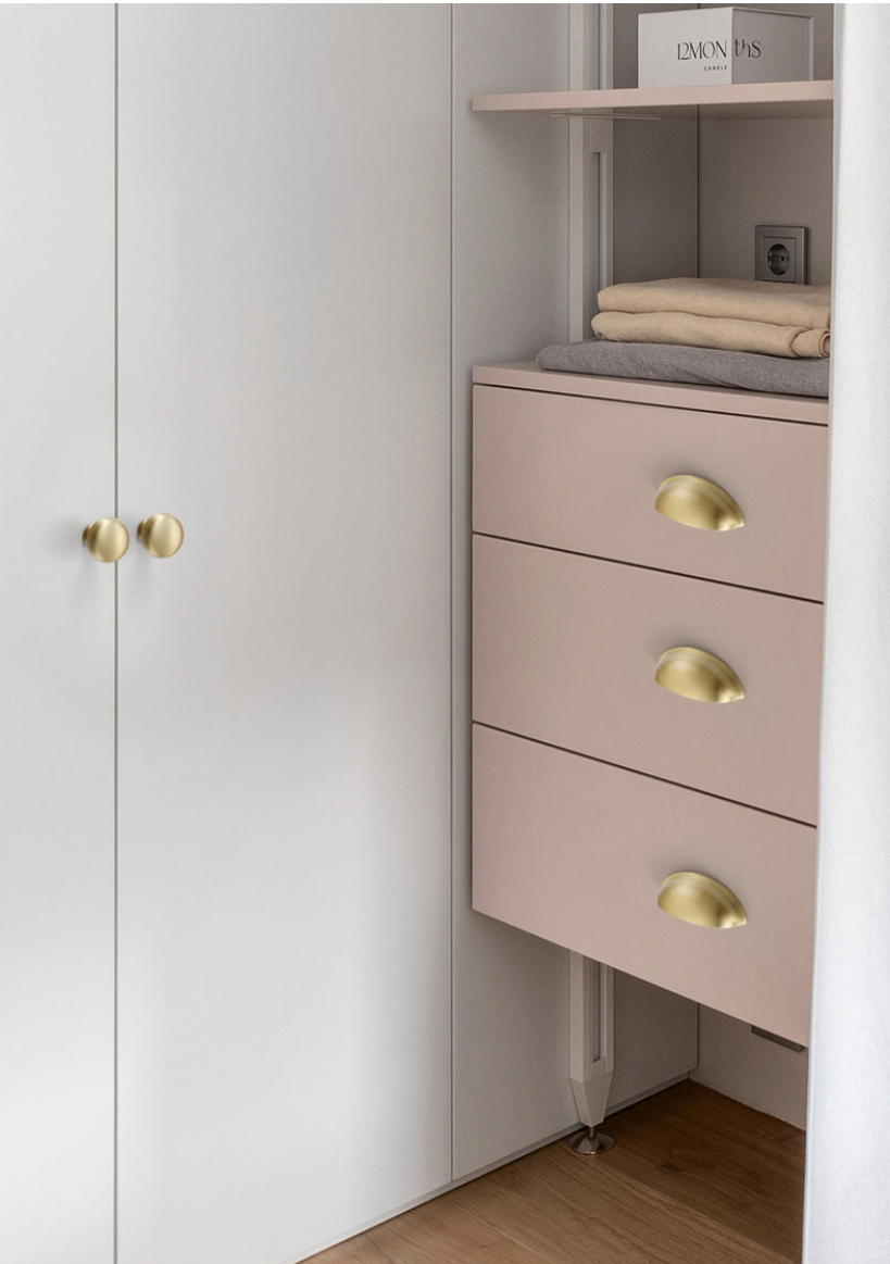 Three satin brass half-moon cup pulls on pale pink drawers beside white cabinet doors.